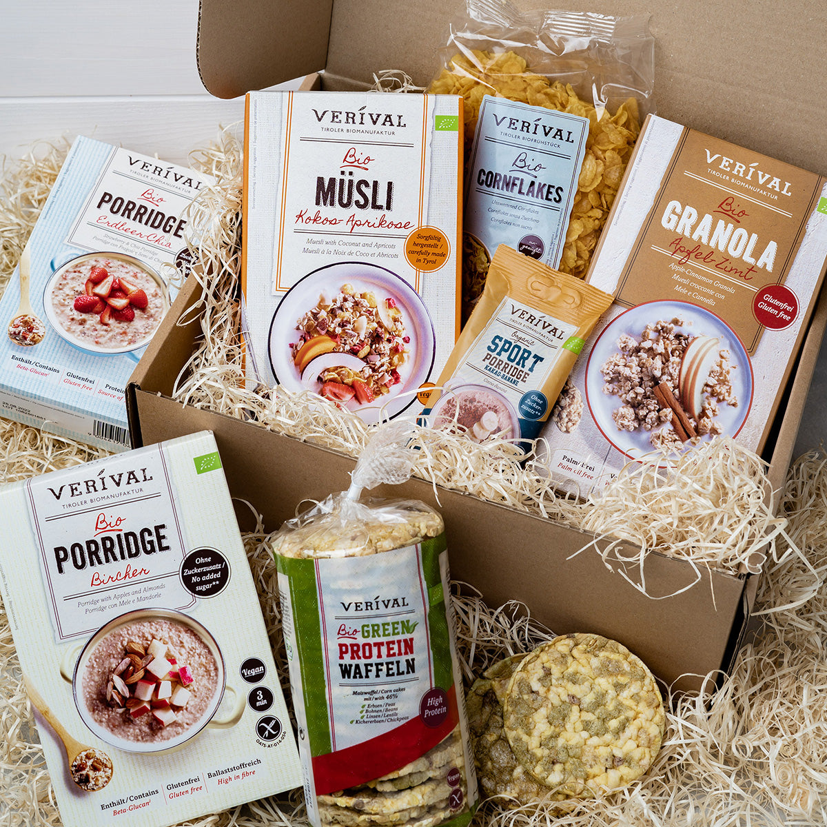Various Verival breakfast products, including porridge, muesli, granola, cornflakes and green protein waffles, presented in a box with straw-like packaging material.