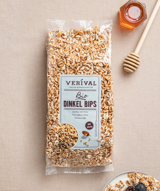 Eine Packung Dinkel Bips mit Honig von Verival Bio  steht zusammen mit einem Honiglöffel, einem Glas Honig und einem Teller mit Trockenfrüchten auf einem Tisch.