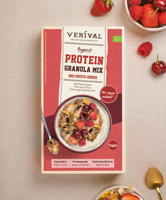 Eine Paket Protein Granola Mix Red Fruits-Kokos von Verival Bio, das ohne Zuckerzusatz und mit veganen pflanzlichen Proteinen aufwartet, wird mit einer Schüssel Granola, umgeben von frischen Früchten und Nüssen, präsentiert.