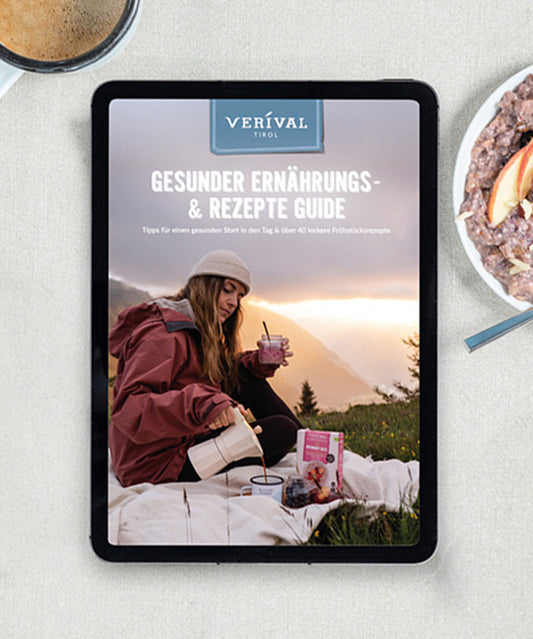 Auf einem Tablet ist das Cover des „Kostenloser Ernährungs- und Rezeptguide Deutsch/Englisch“ der Verival Bio GmbH zu sehen, auf dem eine Frau in rosa Jacke zu sehen ist, die ihr Frühstück im Freien genießt, mit Bergen im Hintergrund. Eine Schüssel Beerenbrei und ein Löffel stehen für nahrhafte Frühstücksrezepte.