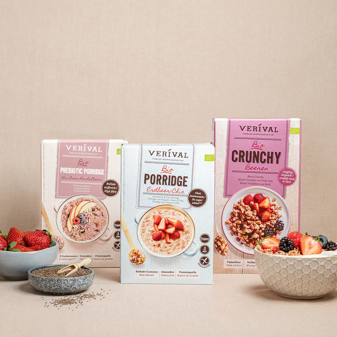 On a beige surface are three Verival cereal boxes with bowls of mixed berries and ready-made muesli, along with a small bowl of chia seeds and a spoon.