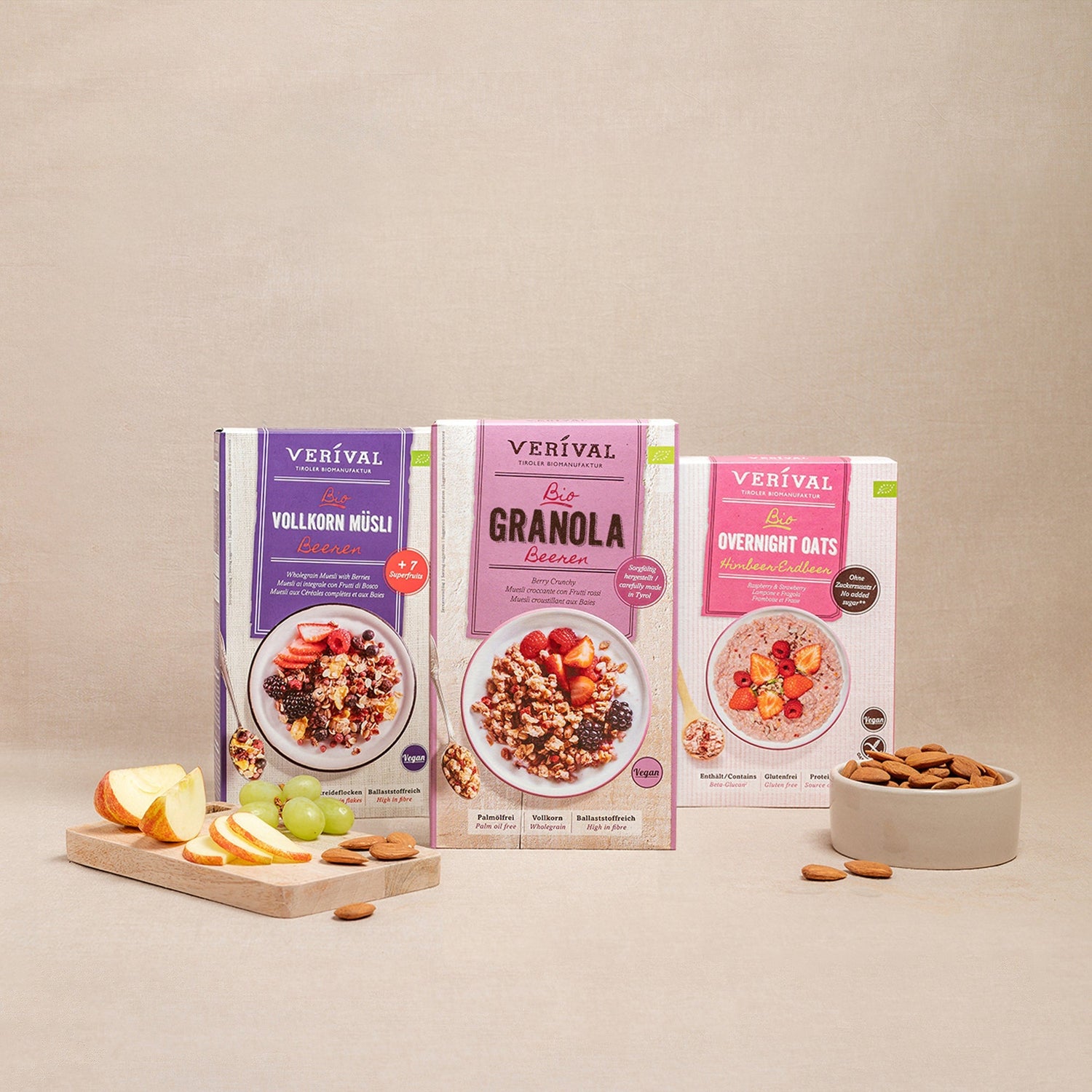 Three packs of Verival muesli (wholemeal muesli, granola, overnight oats) are presented with a bowl of almonds and a cutting board with apple and grape slices.