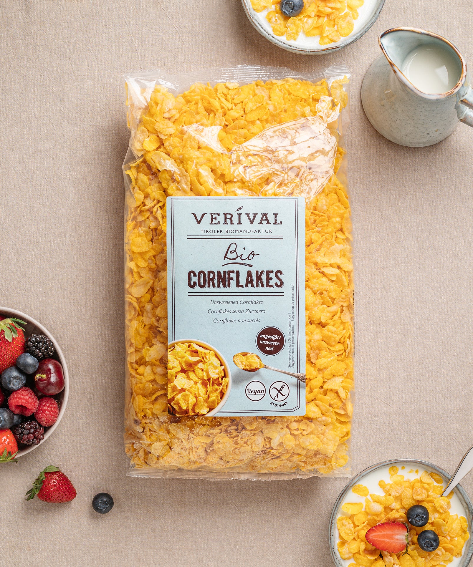 Eine Packung Cornflakes ungesüßt von Verival Bio umgeben von Schalen mit Cornflakes mit Milch und einer Schale mit gemischten Beeren auf einer beigen Tischdecke.