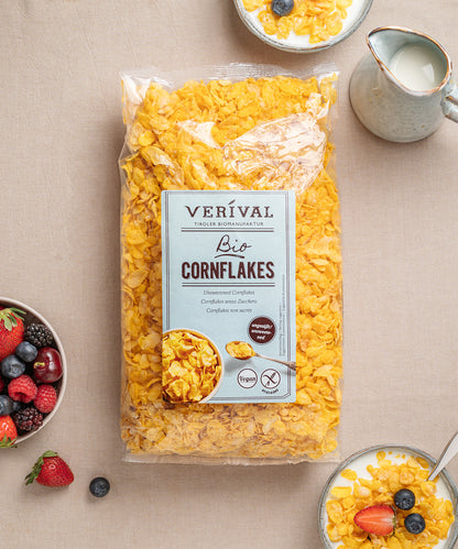 Eine Packung Cornflakes ungesüßt von Verival Bio umgeben von Schalen mit Cornflakes mit Milch und einer Schale mit gemischten Beeren auf einer beigen Tischdecke.