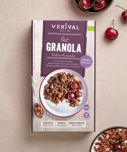 Eine Schachtel Verival Bio GmbH Bio Kakao-Kirsche Granola glutenfrei steht auf einer hellen Fläche, umgeben von Kirschen und einer Schale veganes, glutenfreies Knuspermüsli mit Kirschen.