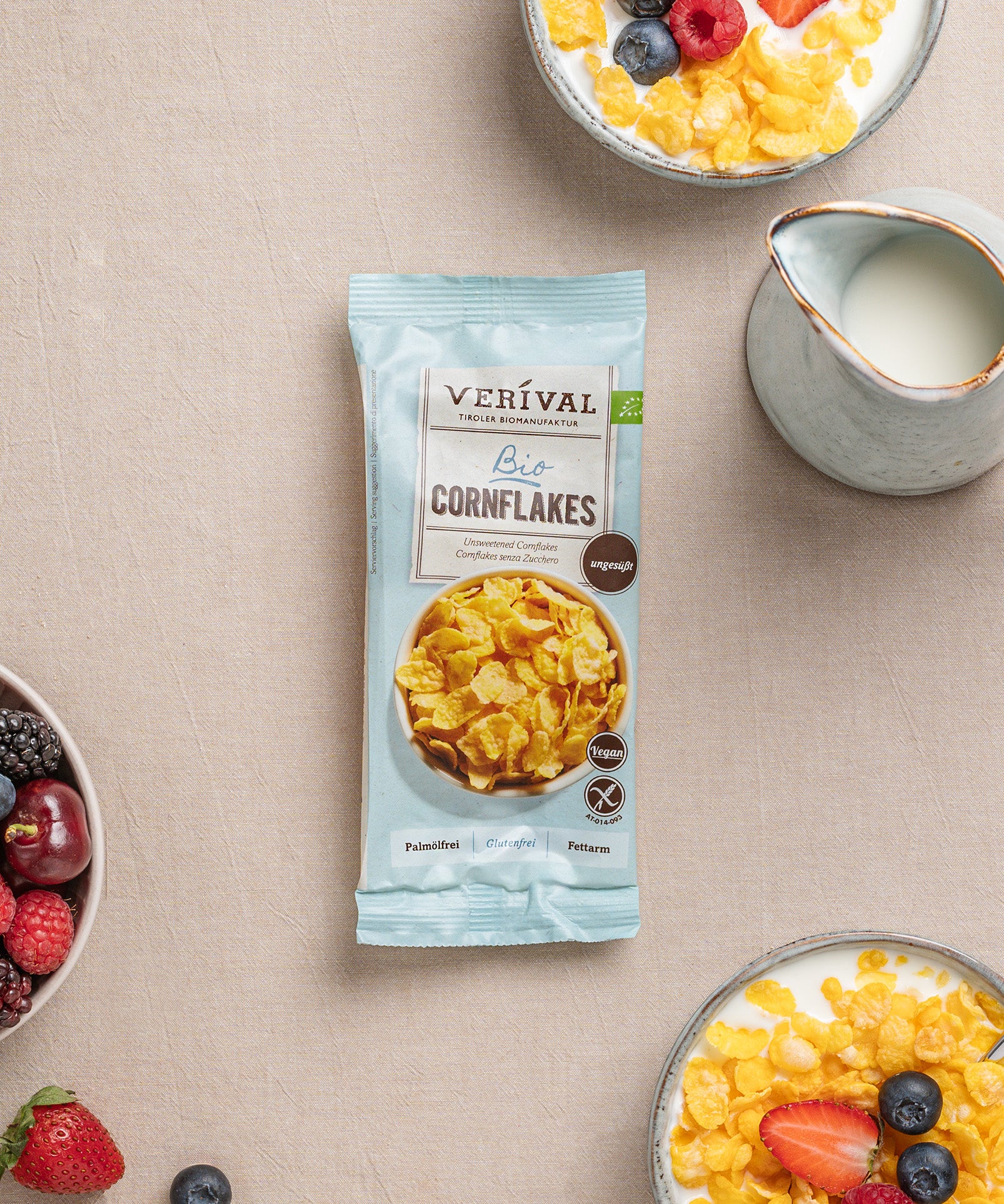 Auf einem Tisch steht eine Einzelportionspackung ungesüßte Cornflakes knusprige Frühstücksoption, daneben Schüsseln mit diesen knusprigen Leckereien, Milch und frischen Beeren.