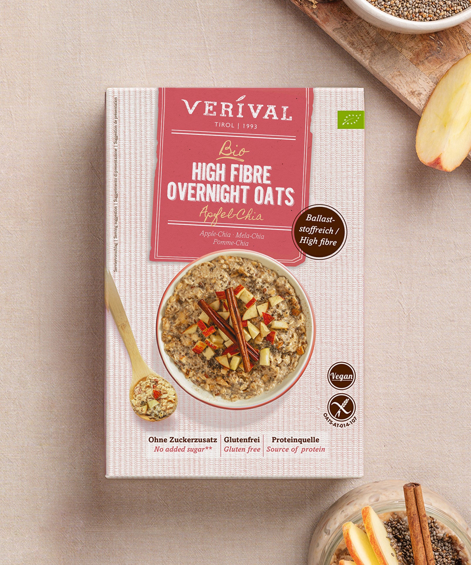 Eine Packung Verival Bio GmbH Bio High Fibre Oats Apfel-Chia steht auf einer beigen Unterlage mit Haferflocken, einer Apfelscheibe und einer Schale mit veganen Haferflocken und Chia - ideal für ein ballaststoffreiches Frühstück.