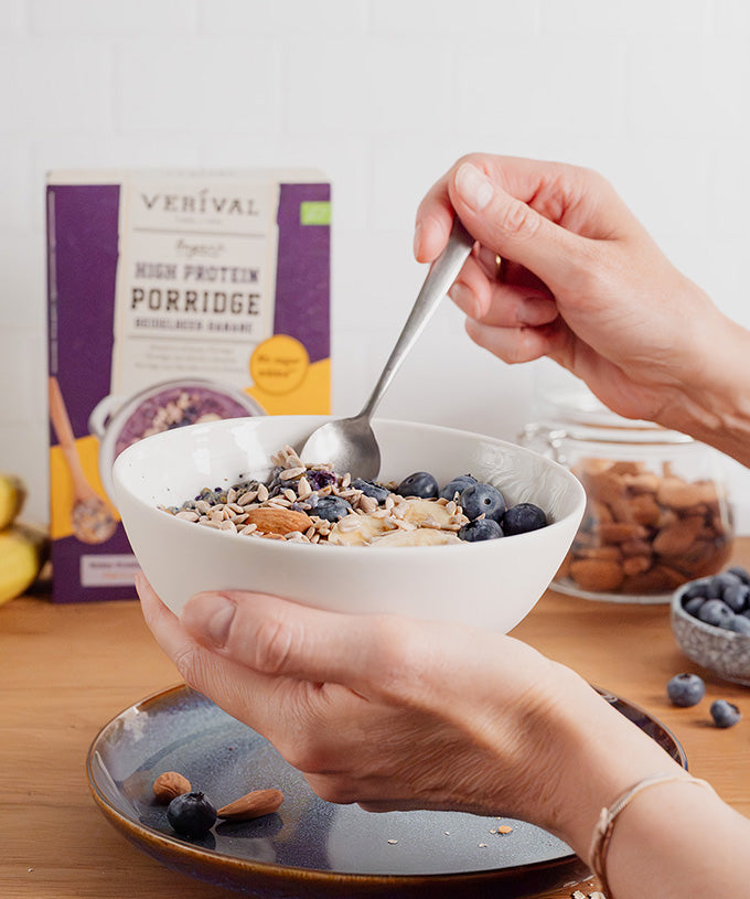 Jemand genießt den „Bio High Protein Porridge Heidelbeere-Banane“ von Verival Bio GmbH, garniert mit Heidelbeeren und Samen. Im Hintergrund sind die Porridge-Packung, ein Glas Mandeln und ein Teller mit Nüssen zu sehen.