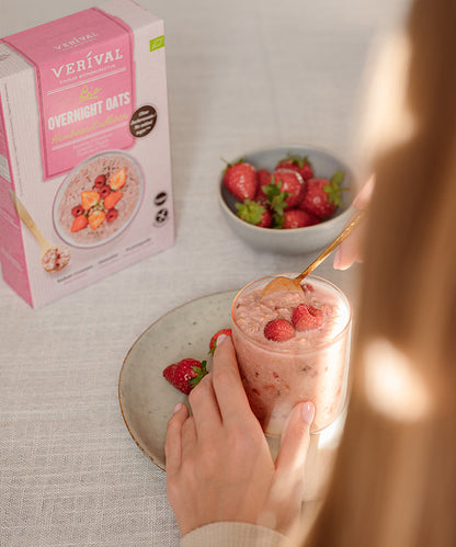Eine Person hält ein Glas „Bio Overnight Oats Himbeer-Erdbeer“ von Verival Bio GmbH in der Hand, daneben steht ein Teller mit einer Erdbeere. Auf dem Tisch stehen eine Schale Erdbeeren und die Produktverpackung – ideal für ein gesundes Frühstück ohne Zuckerzusatz.