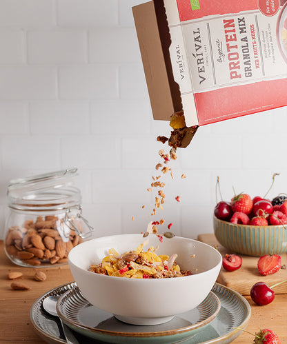 Eine Hand schüttet den „Bio Protein Granola Mix Rote Früchte-Kokos“ der Verival Bio GmbH in eine weiße Schüssel auf einem Tisch. Daneben stehen ein Glas mit Mandeln, eine Schüssel mit Kirschen und Himbeeren sowie ein Löffel auf einem Teller unterhalb der Schüssel.